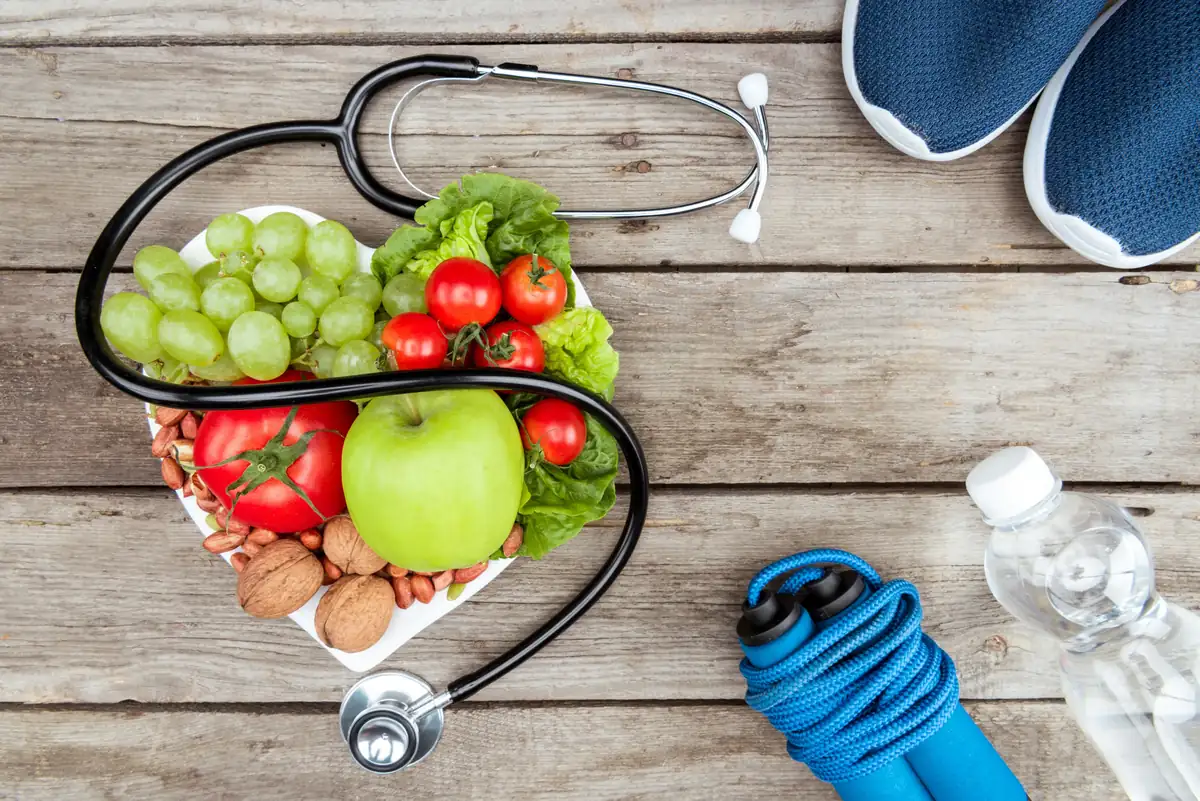 Stethoscope wrapped around a heart-shaped plate of fruit, vegetables and nuts, next to a jump rope, sneakers and water