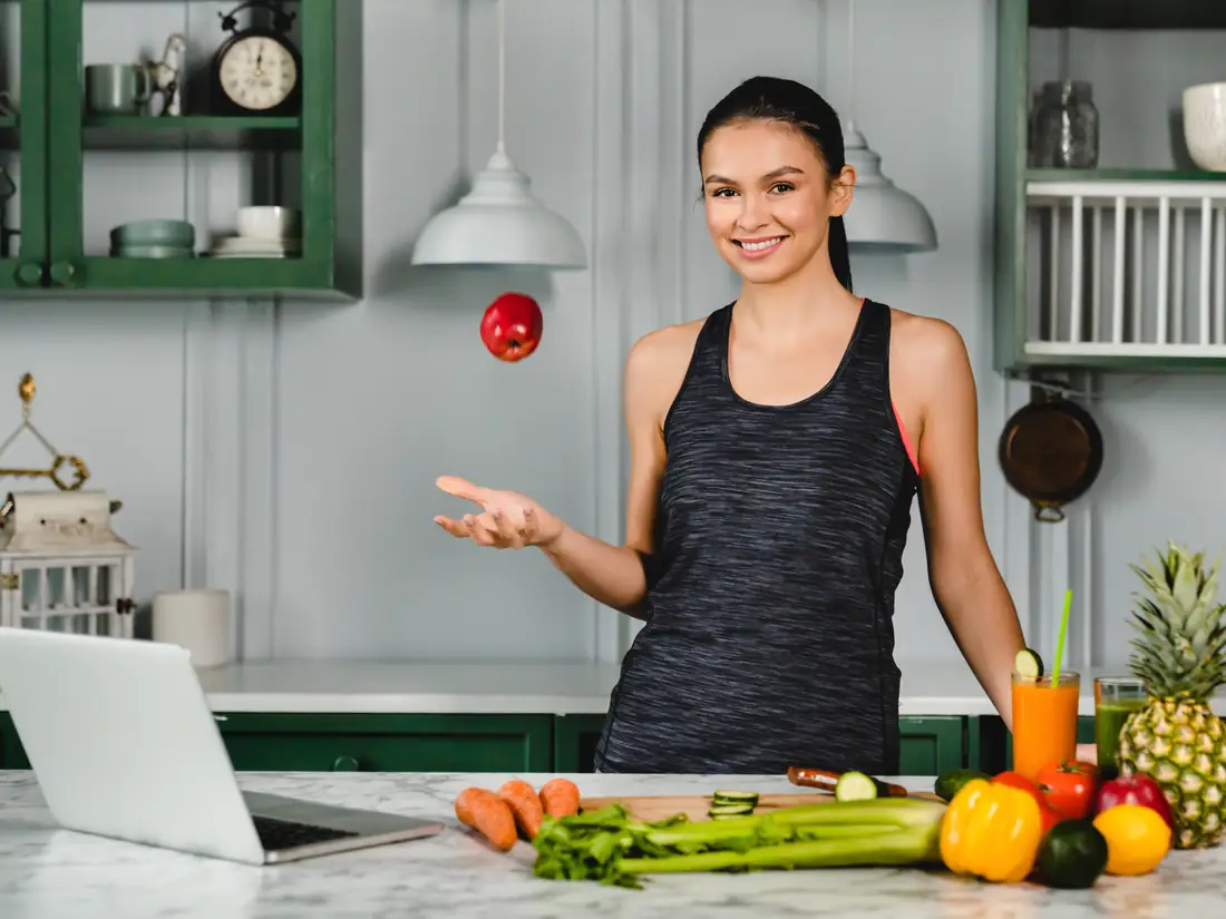 Smiling woman in a bright kitchen with a laptop and fresh vegetables, ready for a virtual nutrition session